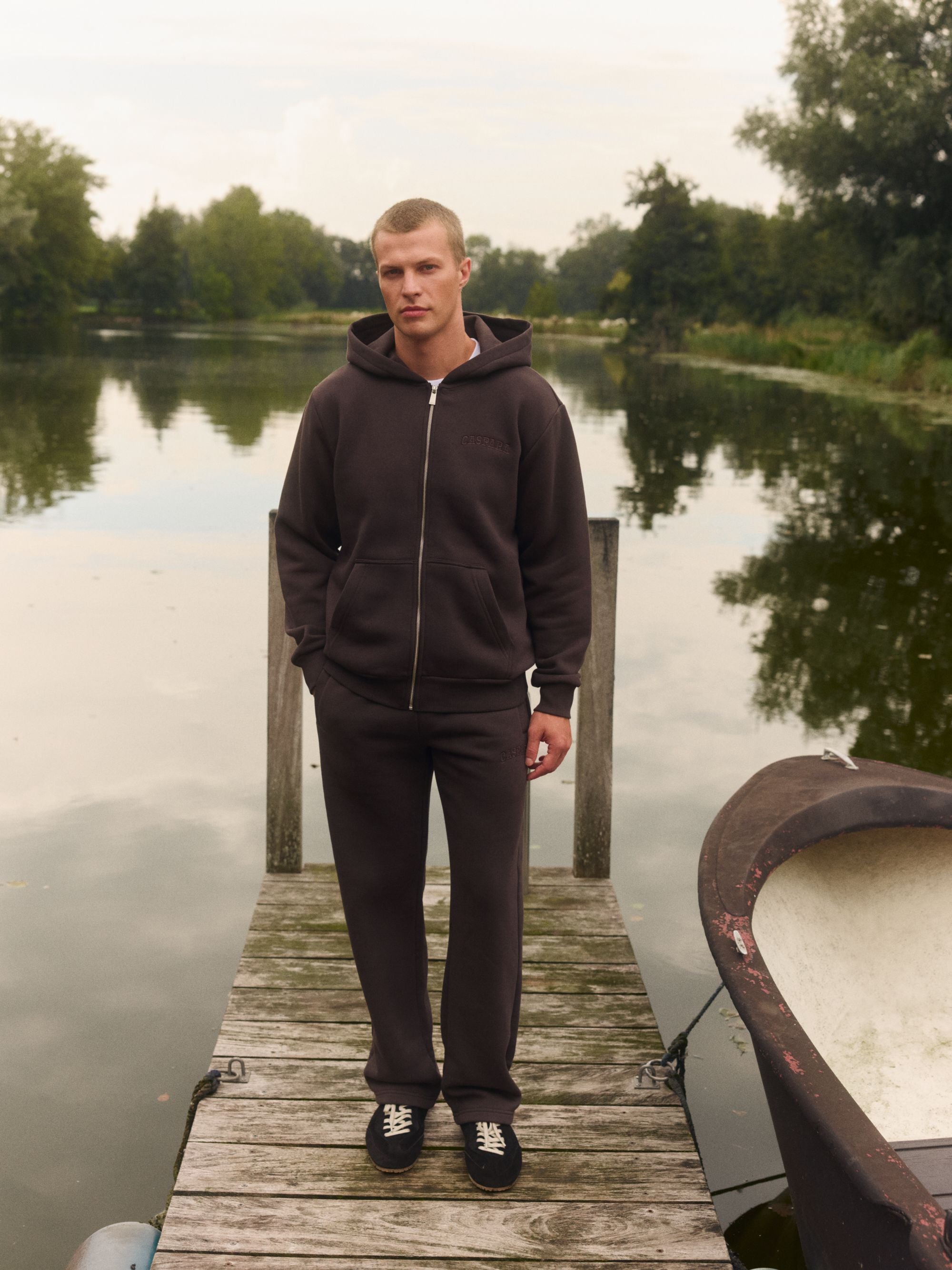 Man wearing an espresso CASPARA zip hoodie and straight-leg pants on a wooden dock by a lake.